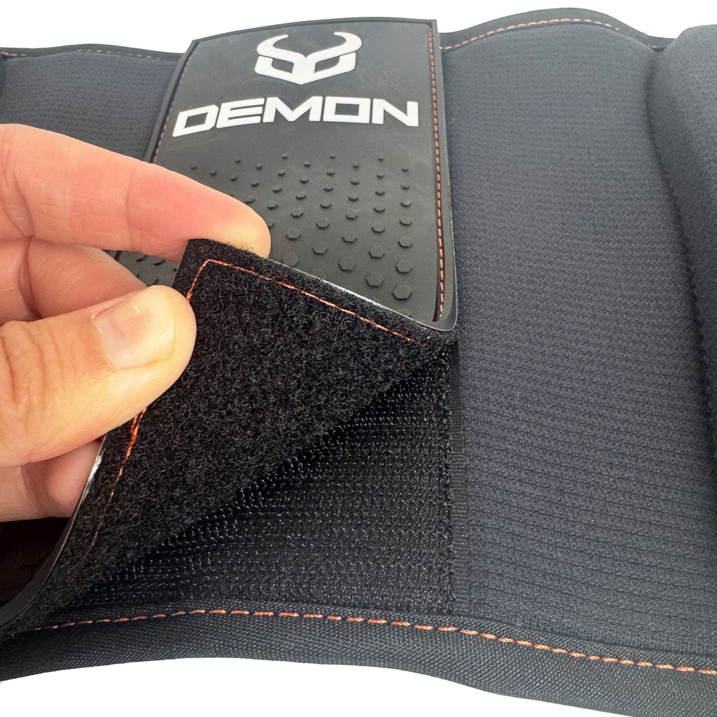 Gray shoulder pad with 'Demon' branding and a hand adjusting the velcro strap.