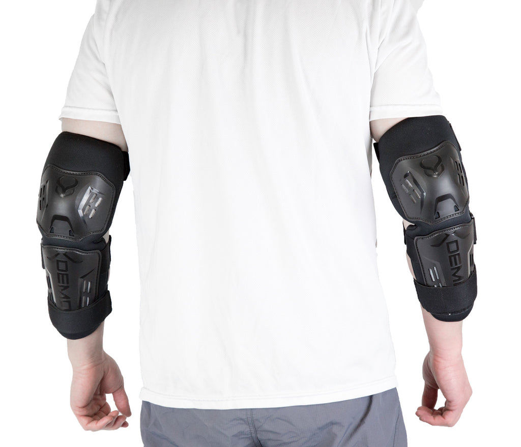 Rear view of a pair of black Demon hard-shell elbow and forearm guards being worn over a white shirt, featuring a dual-plate design and adjustable straps for heavy-duty joint and limb protection.