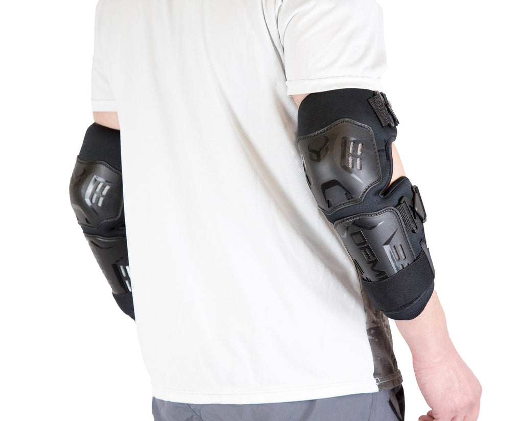 Side view of a pair of black Demon hard-shell elbow and forearm guards being worn over a white shirt, featuring a dual-plate design and adjustable straps for heavy-duty joint and limb protection.